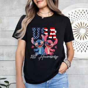 May include: Black t-shirt with a graphic of two cartoon characters wearing sunglasses with the American flag design. The word "USA" is above the characters, and "250th Anniversary" is written below.