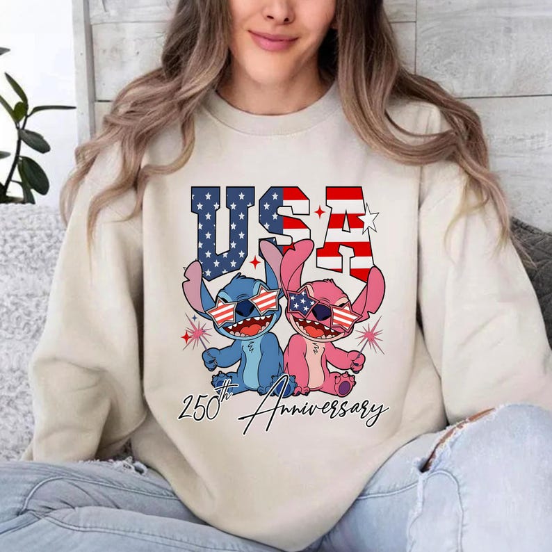 May include: A cream-coloured sweatshirt with a graphic of two cartoon characters wearing sunglasses with American flag designs. The word "USA" is above the characters, and "250th Anniversary" is written below.