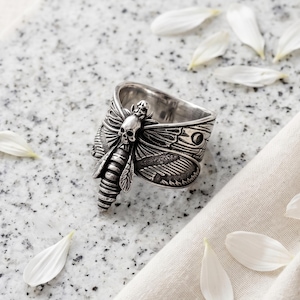 May include: A silver-toned metal ring featuring a detailed moth design with a small skull centered on the thorax. The wings are textured with intricate vein patterns and the body is segmented, showcasing a polished metallic finish throughout.