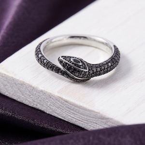 Peut inclure: Une bague argentée en forme de serpent enroulé, dotée d'une surface texturée recouverte de petites pierres noires sombres. La tête du serpent est ornée d'une pierre noire allongée plus grande au centre, offrant un aspect métallique élégant.