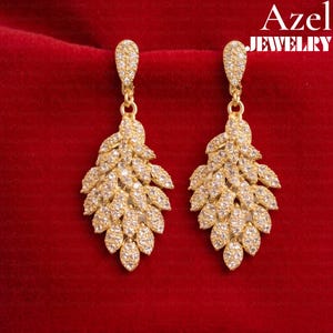 May include: A pair of gold-toned drop earrings featuring a leaf-shaped design encrusted with numerous small, clear, sparkling stones. Each earring has a teardrop-shaped top piece connected to a larger, multi-layered leaf cluster. Text reads Azel JEWELRY.