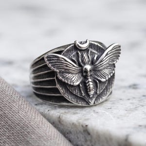 May include: A silver-toned ring featuring a detailed moth design centered on a circular plate. The moth has prominent wings and a skull-like marking on its thorax. A small crescent moon symbol is positioned directly above the moth's head.