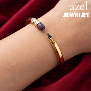 May include: A gold-toned cuff bracelet shaped like a pencil, featuring a purple pavé-set eraser end with clear stones and a black pavé-set tip. The text azel JEWELRY is visible in the upper right corner against a dark red fabric background.