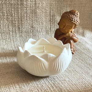 Op de afbeelding: Een witte kaarshouder in de vorm van een lotusbloem met een crèmekleurige waskaars met een enkele centrale lont, geplaatst naast een klein, bruin, zittend Boeddhabeeldje met een textuurrijk oppervlak en gesloten ogen.