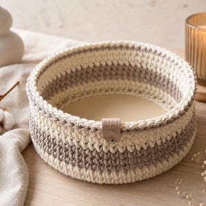 May include: A round, hand-crocheted basket featuring alternating horizontal stripes of cream and light brown yarn. The basket has a smooth, solid base and a small, rectangular beige leather tag attached to the rim with the text Made with LOVE.