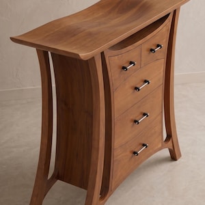 Puede incluir: Una cómoda de madera con una parte superior curvada y cuatro cajones. La cómoda está hecha de madera de color marrón claro y tiene un diseño único y moderno. Los cajones tienen tiradores plateados. Las patas son curvas e inclinadas, lo que añade un aspecto distintivo.