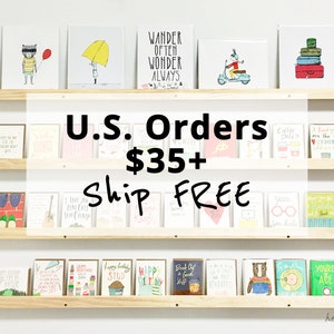 May include: A shelf display of greeting cards with various illustrations and text. The cards are arranged in three rows. The text on the sign reads "U.S. Orders $35+ Ship FREE".