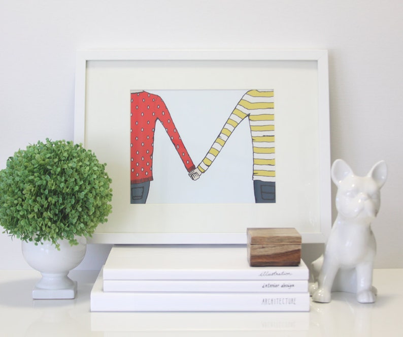 May include: A white framed illustration of two people holding hands, forming the letter "M". The person on the left is wearing a red sweater with white polka dots, and the person on the right is wearing a yellow and white striped sweater. The illustration is on a white background.