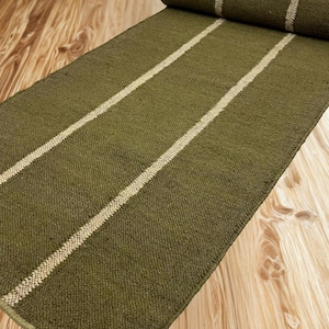 May include: A rectangular olive green runner rug featuring two parallel cream-colored stripes running the length of the fabric. The rug has a textured, woven appearance and is partially rolled at one end, displaying a consistent weave pattern.