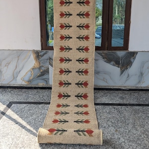 May include: A long, rectangular runner rug made of natural tan jute fiber. The rug features a repeating pattern of stylized floral motifs arranged in two vertical columns. Each motif consists of a red petal shape above dark green leaves.