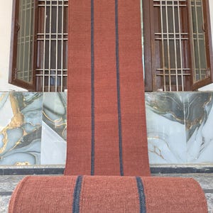 Puede incluir: Un camino de mesa rectangular largo de tela texturizada color terracota, con dos franjas verticales distintivas en gris carbón oscuro que recorren toda la pieza. El camino está parcialmente enrollado en la base, mostrando un tejido uniforme.