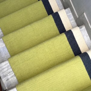 Puede incluir: Una serie de alfombrillas rectangulares para escaleras con un centro texturizado en verde lima, una franja sólida en gris oscuro y un borde de color crema. El tejido cubre la superficie horizontal de cada escalón, creando un patrón.