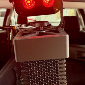 Könnte beinhalten: Ein schwarzer Bühnenscheinwerfer mit quadratischem Kopf und vier kreisförmigen roten LED-Leuchten. Das Gerät ist auf einer Halterung mit zentralem Einstellknopf montiert und verfügt über einen hohen, vertikalen Sockel mit schwarzem Wabengitter.