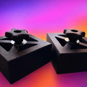 Puede incluir: Dos bloques geométricos negros con perillas en forma de estrella. Cada bloque tiene un recorte triangular y una arandela plateada debajo de la perilla. El fondo es un degradado de morado, naranja y amarillo.