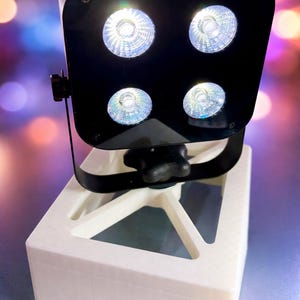 Könnte beinhalten: Ein schwarzer, quadratischer LED-Strahler mit vier kreisförmigen Linsen, die hellweißes Licht ausstrahlen, montiert auf einer schwarzen, verstellbaren Halterung auf einem weißen 3D-gedruckten Sockel mit dem Schriftzug northshore sound.