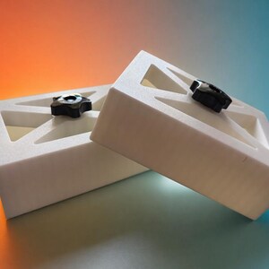 May include: Two white rectangular blocks with a hollowed-out geometric design on the top surface. Each block features a black star-shaped knob centered within the cutout. The blocks are positioned at an angle, showing their solid white sides.