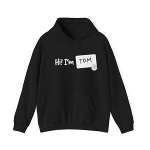 May include: Black hooded sweatshirt with a front pocket and drawstring. The text "Hi! I'm TOM" is printed in white, along with a name tag graphic. A small graphic of a clock is on the name tag.