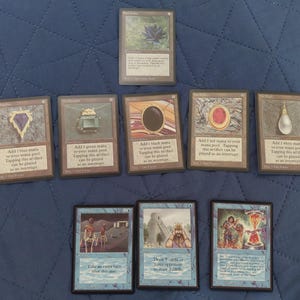 Puede incluir: Una colección de cartas coleccionables de Magic: The Gathering, con varios artefactos y hechizos. Las cartas muestran ilustraciones coloridas y texto, con un enfoque en los símbolos de maná y la mecánica del juego. Las cartas están dispuestas sobre una superficie azul.