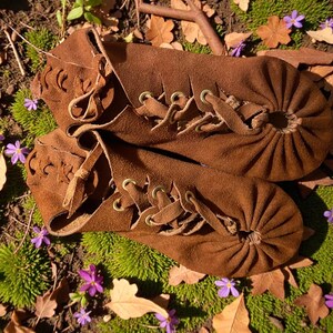 May include: A pair of brown suede moccasins with lace-up detailing. The shoes have a gathered toe design and decorative cutouts. The image is set outdoors, with the shoes resting on a bed of green moss, fallen leaves, and small purple flowers.
