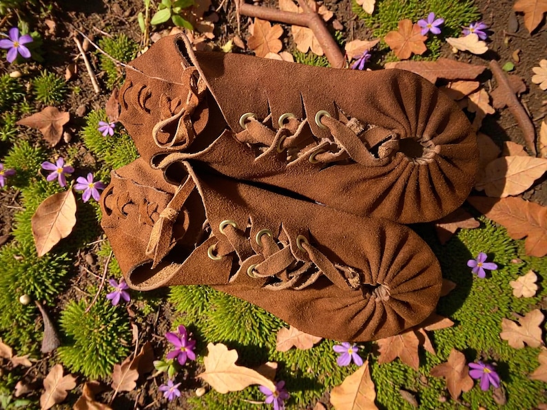 May include: A pair of brown suede moccasins with lace-up detailing. The shoes have a gathered toe design and decorative cutouts. The image is set outdoors, with the shoes resting on a bed of green moss, fallen leaves, and small purple flowers.