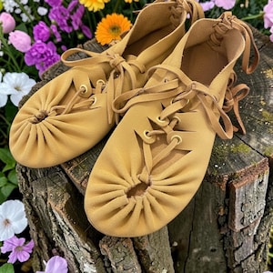 May include: A pair of tan leather moccasin-style shoes with decorative stitching and laces. The shoes are displayed on a tree stump, surrounded by colorful flowers. The shoes have a soft, suede-like texture.