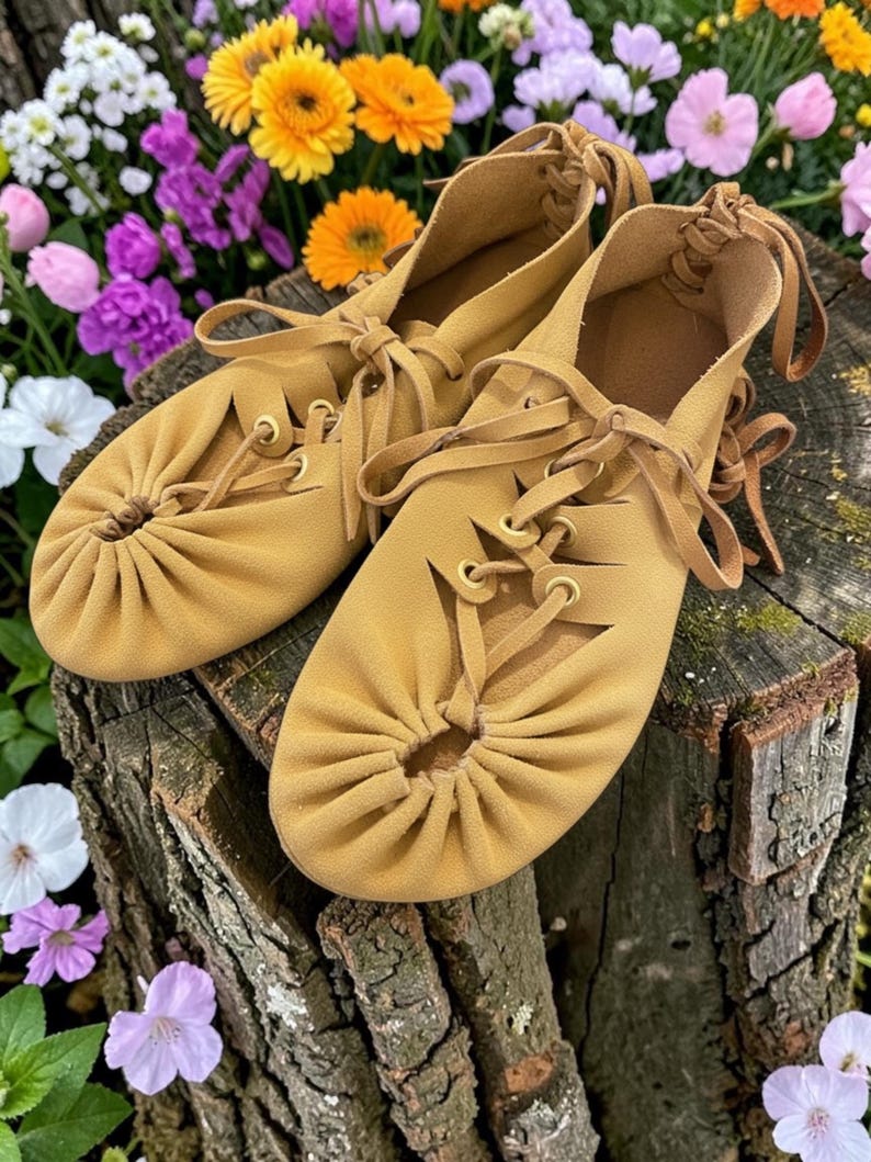May include: A pair of tan leather moccasin-style shoes with decorative stitching and laces. The shoes are displayed on a tree stump, surrounded by colorful flowers. The shoes have a soft, suede-like texture.