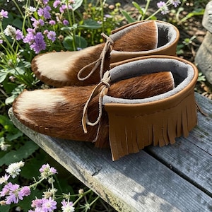 Puede incluir: Un par de mocasines confeccionados en piel de vaca marr&oacute;n y blanca con un acabado de pelo texturizado. El calzado presenta un detalle de flecos de cuero color canela alrededor del cuello, forro interior gris suave y cordones de cuero finos.