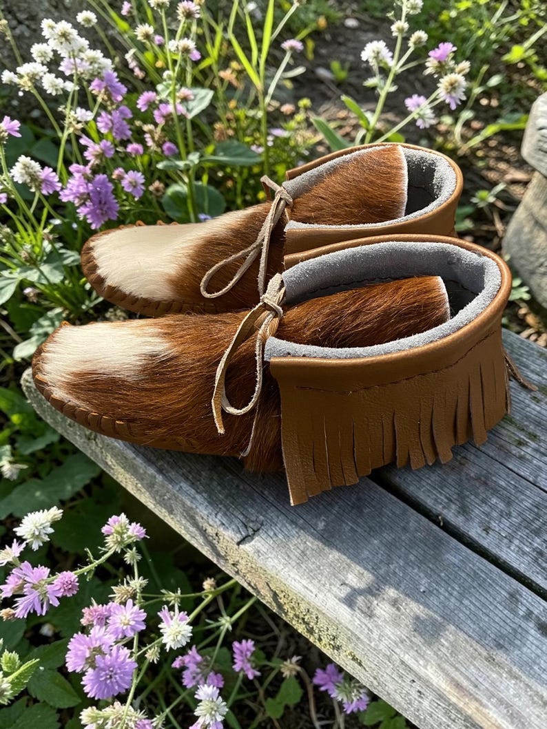 Puede incluir: Un par de mocasines confeccionados en piel de vaca marr&oacute;n y blanca con un acabado de pelo texturizado. El calzado presenta un detalle de flecos de cuero color canela alrededor del cuello, forro interior gris suave y cordones de cuero finos.