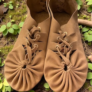 May include: A pair of brown leather moccasin-style shoes with laces. The shoes have a gathered design at the toe and are set against a backdrop of green foliage and moss.