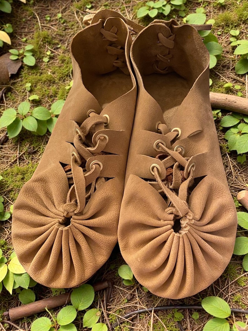 May include: A pair of brown leather moccasin-style shoes with laces. The shoes have a gathered design at the toe and are set against a backdrop of green foliage and moss.