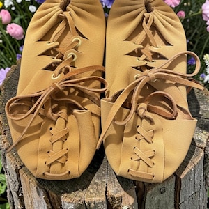 May include: A pair of tan leather moccasin-style slippers with lace-up detailing. The slippers have a soft, rounded toe and are tied with leather laces. The background features colorful flowers.
