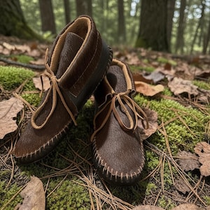 May include: A pair of brown leather moccasin boots with tan laces and stitching. The boots have a dark brown fur-like texture and are set against a backdrop of moss, leaves, and pine needles.