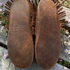 Puede incluir: Un par de mocasines de cuero marr&oacute;n con detalles de flecos visibles en los laterales. Las suelas son planas y est&aacute;n construidas con una sola pieza de cuero marr&oacute;n texturizado, mostrando vetas naturales y costuras en los bordes exteriores.