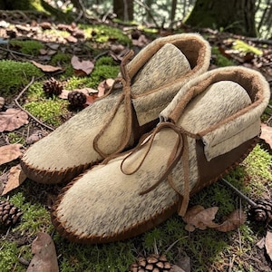 May include: A pair of handmade moccasin-style boots. The boots feature a light-colored, textured upper with brown leather accents and stitching. They have a fur-like lining and are laced with leather ties. The boots are set on a forest floor with moss and pinecones.