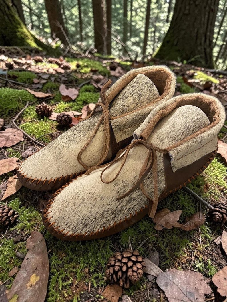 May include: A pair of handmade moccasin-style boots. The boots feature a light-colored, textured upper with brown leather accents and stitching. They have a fur-like lining and are laced with leather ties. The boots are set on a forest floor with moss and pinecones.