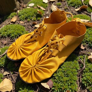 May include: A pair of bright yellow leather shoes with laces. The shoes have a gathered design on the toe and are set on a bed of green moss and fallen leaves. The laces are tied at the top.