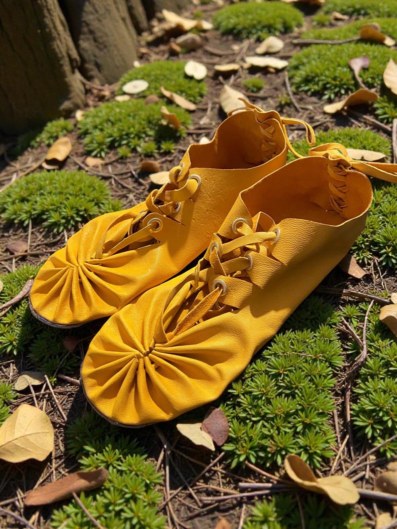 May include: A pair of bright yellow leather shoes with laces. The shoes have a gathered design on the toe and are set on a bed of green moss and fallen leaves. The laces are tied at the top.