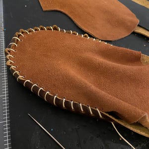 May include: Close-up of leather craft in progress. Two pieces of brown leather, one with a stitched edge using white thread. A needle and thread are also visible, suggesting a handmade item.