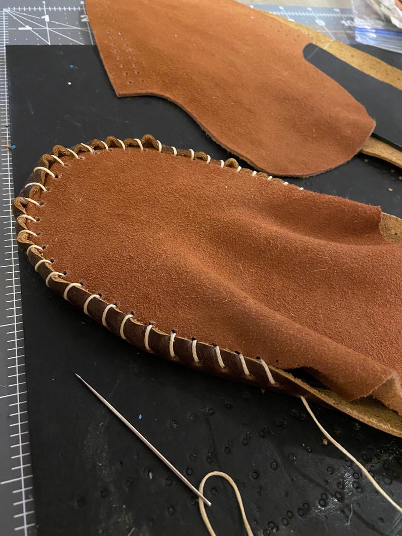 May include: Close-up of leather craft in progress. Two pieces of brown leather, one with a stitched edge using white thread. A needle and thread are also visible, suggesting a handmade item.