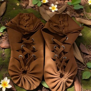 May include: A pair of brown leather moccasin-style shoes with lace-up detailing. The shoes have a gathered toe design and are set against a natural backdrop of moss, leaves, and flowers. The footwear appears handmade.