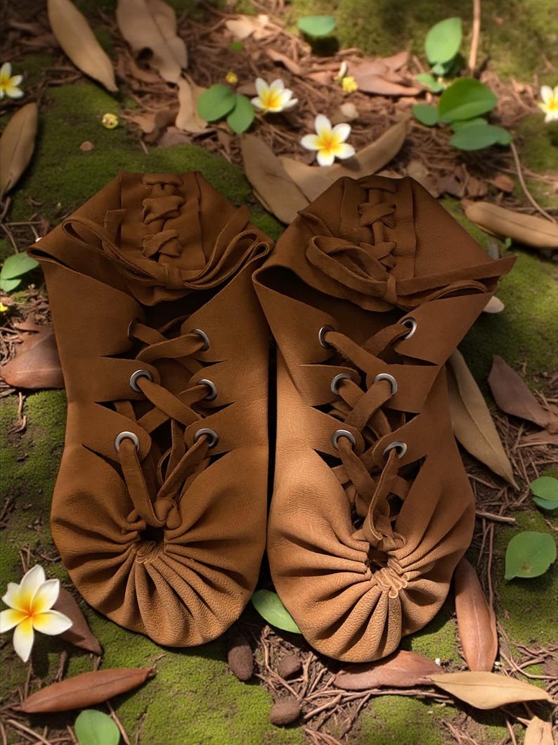 May include: A pair of brown leather moccasin-style shoes with lace-up detailing. The shoes have a gathered toe design and are set against a natural backdrop of moss, leaves, and flowers. The footwear appears handmade.