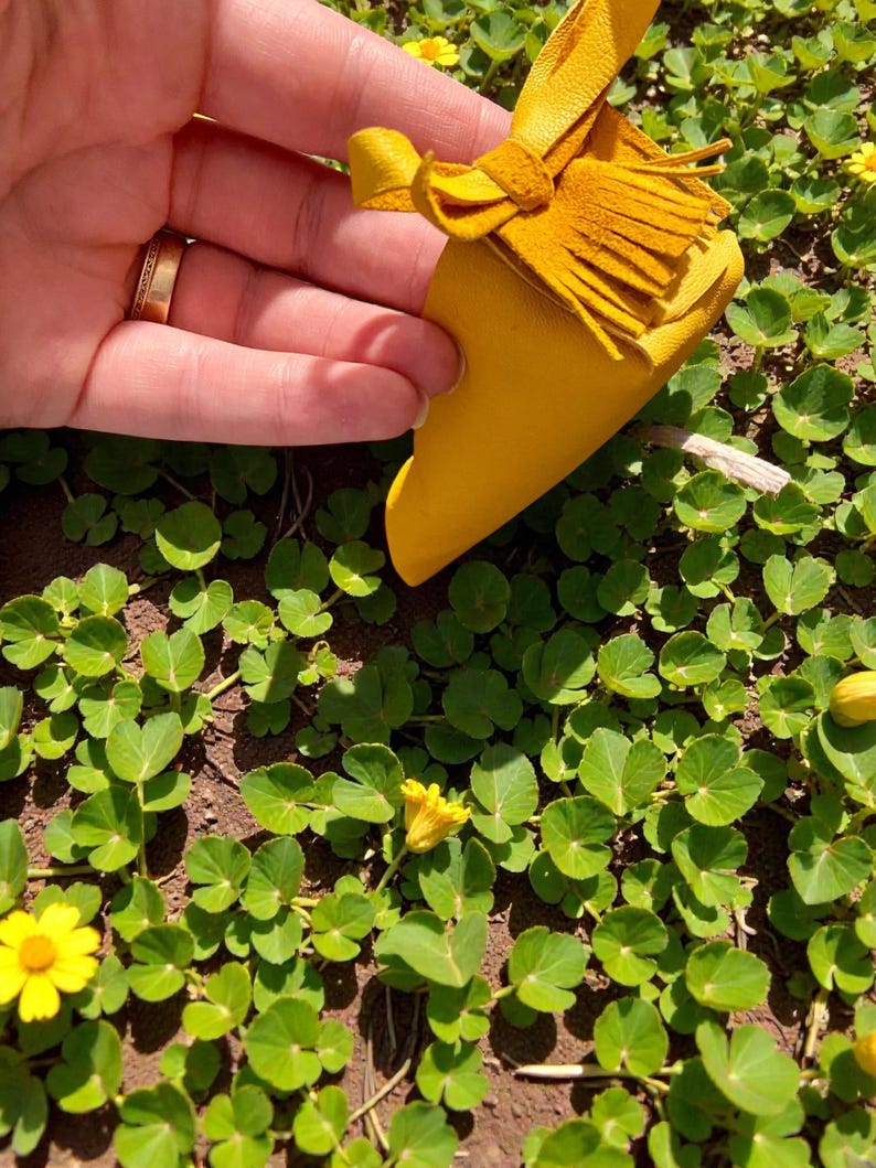 May include: A small, yellow leather pouch with a tassel and bow detail. The pouch is held against a backdrop of green foliage and small yellow flowers, suggesting a natural setting. The leather appears to be soft and the pouch is likely handmade.