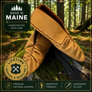 May include: Tan leather moccasins featuring visible dark brown stitching along the seams and a soft, flexible construction. The footwear includes text labels stating Made in Maine, Handcrafted with Care, Lifetime Repairs & Resoling, Premium Natural Leather, Barefoot Friendly, and Handmade to Last.