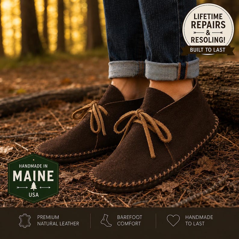 May include: Dark brown suede leather moccasin boots with tan leather laces and visible whipstitch detailing along the sole. The boots are worn with cuffed blue jeans. Text overlays include "LIFETIME REPAIRS & RESOLING!" and "HANDMADE IN MAINE USA".