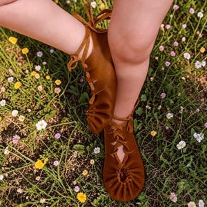 May include: Brown leather moccasins with lace-up detailing. The shoes are shown on a person's feet, surrounded by a field of green grass and wildflowers. The moccasins have a soft, flexible sole and are tied with leather laces.
