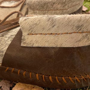 May include: Close-up of a pair of handmade moccasins. The shoes feature a combination of brown leather and light-colored fur. The edges are stitched with orange thread, and the laces are a light brown leather. The moccasins are designed for comfort and style.