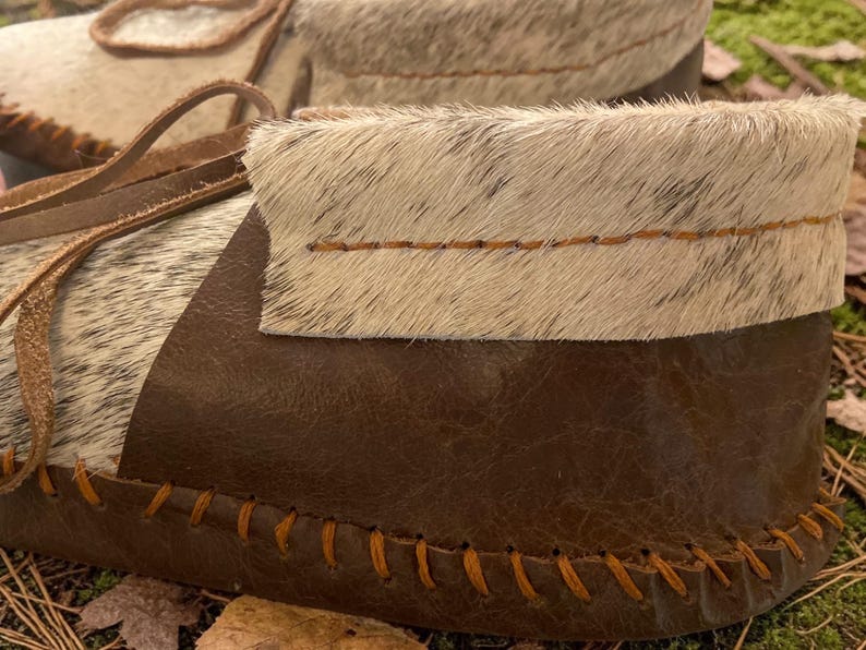 May include: Close-up of a pair of handmade moccasins. The shoes feature a combination of brown leather and light-colored fur. The edges are stitched with orange thread, and the laces are a light brown leather. The moccasins are designed for comfort and style.