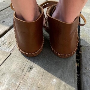 May include: A pair of brown leather moccasins featuring a visible whipstitch detail along the sole edge. The footwear has a low-cut ankle design with leather laces visible at the sides, showcasing a smooth, natural grain texture on the leather surface.