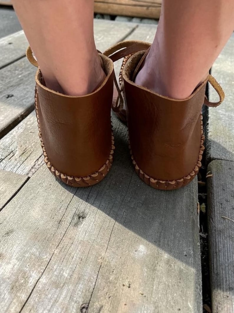 May include: A pair of brown leather moccasins featuring a visible whipstitch detail along the sole edge. The footwear has a low-cut ankle design with leather laces visible at the sides, showcasing a smooth, natural grain texture on the leather surface.