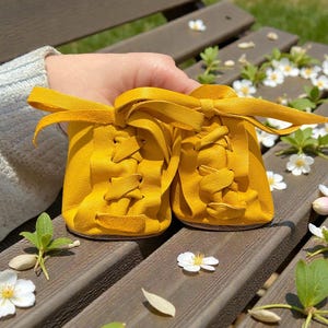 May include: A pair of bright yellow baby shoes with laces and a bow. The shoes are made of soft leather and are sitting on a wooden bench. The background includes white flowers and greenery.